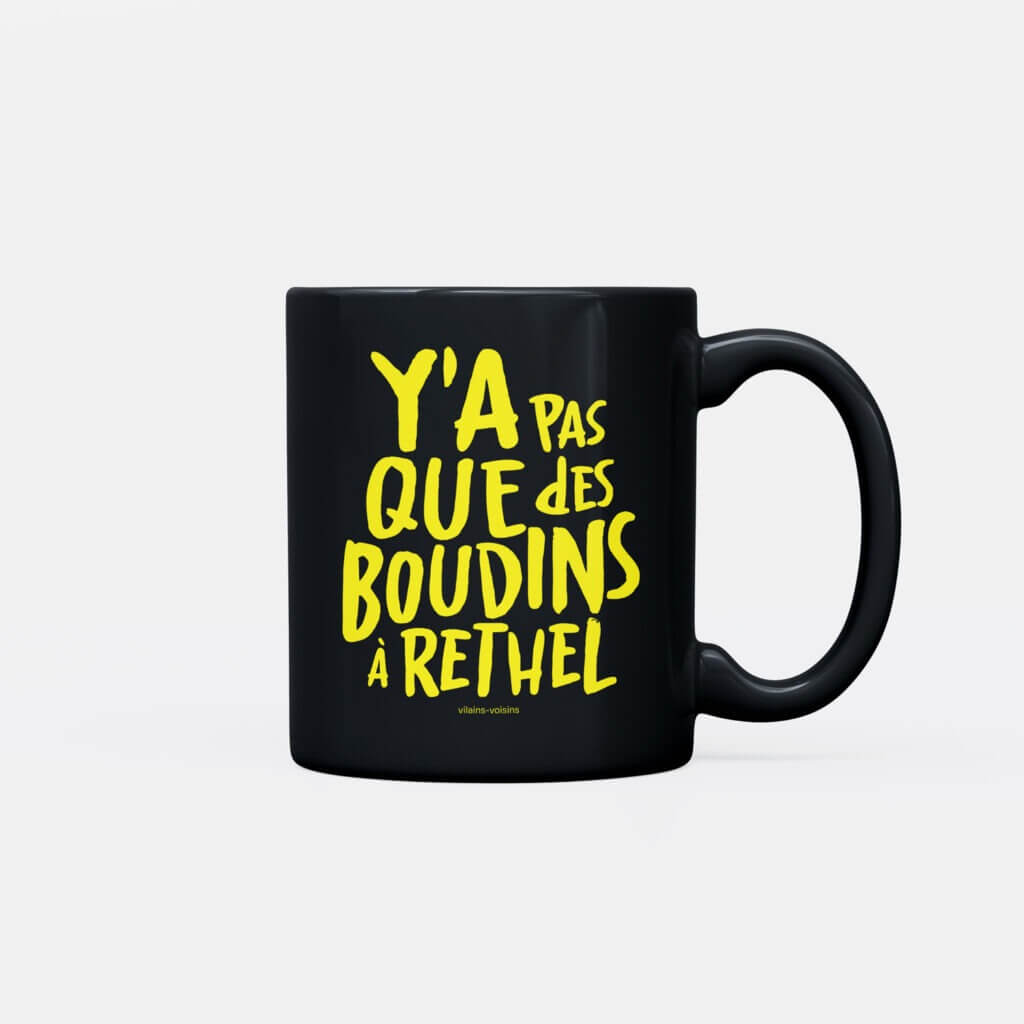 Mug noir, avec le texte y'a pas que des boudins à Rethel en jaune, de vilains-voisins imprimerie et studio de design graphique à Rethel, dans les Ardennes.